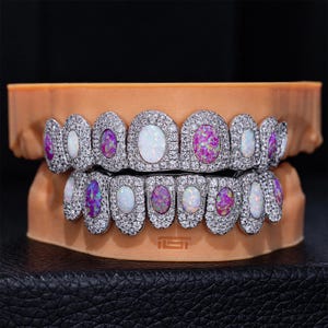 May include: A set of dental grills featuring two rows of teeth covers. Each tooth is encrusted with small clear stones and features a central oval-shaped inlay of either iridescent white or vibrant purple opal-like material, set on a model.