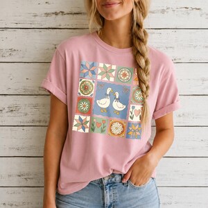 Puede incluir: Una camiseta de manga corta en color rosa empolvado con un estampado gráfico central de dos ocas blancas con lazos azules, rodeadas por una cuadrícula de cuadrados estilo colcha con motivos florales, estrellas, corazones y lazos en tonos pastel.