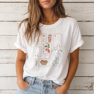 Könnte beinhalten: Ein weißes Kurzarm-T-Shirt mit einem Grafikdruck aus mehreren rechteckigen und ovalen Rahmen, die Illustrationen von Gänsen, Erdbeeren, einem Korb mit Äpfeln, Tomaten, einer Teekanne und blauen Blumen enthalten.