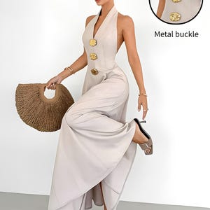 May include: A beige sleeveless halter-neck maxi dress featuring a deep V-neckline and three large, textured gold-tone metal buttons down the front. The dress has an open back design and is paired with a brown woven handbag and grey heeled sandals.
