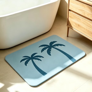 May include: A rectangular, light blue bath mat featuring two dark blue palm tree silhouettes. The mat is placed on a tiled floor near a white bathtub and wooden cabinet, creating a tropical bathroom theme.