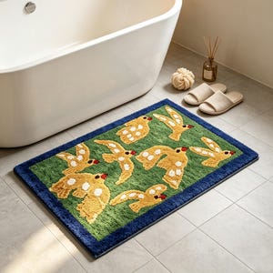 May include: A rectangular bath mat featuring a green background with a pattern of yellow birds with white spots and red beaks, framed by a solid dark blue border. The rug has a textured, tufted surface and is placed on a light gray tiled floor.