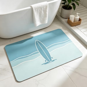May include: A rectangular bath mat with a surfboard design. The mat features a gradient of blue hues, with a white surfboard graphic and white wave lines. The mat is placed on a tiled floor near a white bathtub.