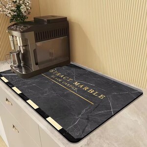 May include: A black marble-patterned kitchen mat with gold text that reads "STRACT MARBLE BLACK CARPET". The mat is rectangular and has a black border with gold accents. A coffee machine sits on the mat.