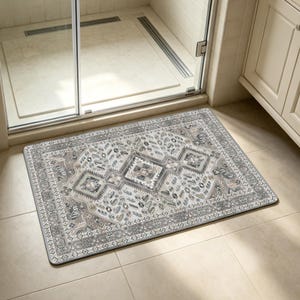 May include: A rectangular, patterned bath mat with a traditional rug design. The mat features a central geometric pattern in shades of gray, beige, and white, with a detailed border. It is placed in front of a shower door.