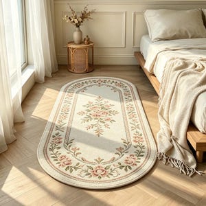 May include: An oval-shaped area rug featuring a cream-colored base with a central floral bouquet and a decorative border of pink roses and green leaves. The rug is positioned on a light wood floor next to a bed with a cream-colored throw blanket.