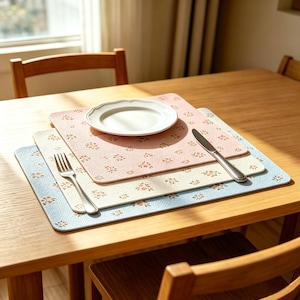May include: A wooden table set with three floral patterned placemats in pink, cream, and light blue. A white plate, a fork, and a knife are on top of the placemats. The placemats have a repeating floral design.