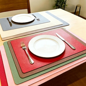 May include: A stack of colorful placemats in shades of red, green, brown, and pink, with a white plate, fork, and knife on top. Additional placemats in gray and beige are visible in the background, each with a plate, fork, and knife.
