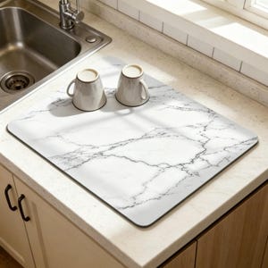 May include: A white and black marble-patterned kitchen mat with two gray mugs. The mat is rectangular with rounded corners and is placed on a white countertop near a stainless steel sink.