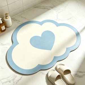 May include: A cloud-shaped bath mat with a light blue border and a heart in the center. The mat is cream-colored inside the border and heart. Two pairs of white sandals are next to the mat. The mat is on a white marble floor.