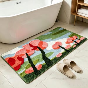 May include: A rectangular bath mat featuring a tufted landscape design with stylized trees in shades of pink, red, and dark green against a light blue and white sky background. The mat has a green border and is paired with a pair of beige open-toe slippers.