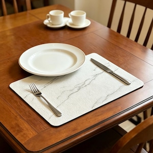 May include: A dining table set with a white marble-patterned placemat, a white plate with a gold rim, a fork, and a knife. Two white teacups and saucers are visible in the background. The table is made of dark wood.