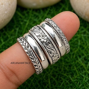 May include: A set of five silver-toned rings stacked together, featuring a mix of smooth polished bands and bands with intricate floral, leaf, and geometric embossed patterns. The text ARCANAMETALS is visible on the side of the ring set.