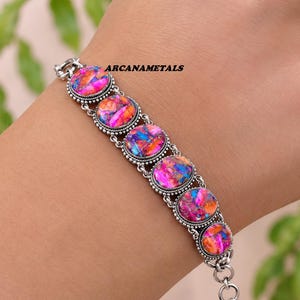 May include: A silver link bracelet featuring six oval-shaped stones with a vibrant, multi-colored pattern of pink, orange, and blue. Each stone is set in a decorative silver bezel with beaded edges. The text ARCANAMETALS is visible on the bracelet.