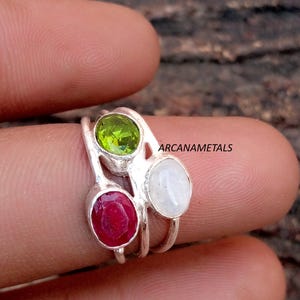 May include: A silver ring featuring three oval-shaped gemstones: a vibrant green peridot, a deep red ruby, and a milky white moonstone, each set in a simple metal bezel. The text ARCANAMETALS is visible near the stones on the silver band.