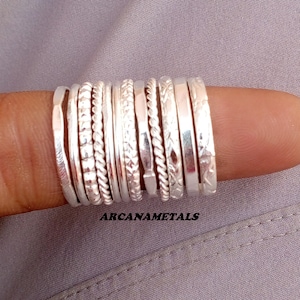 May include: A set of ten silver-coloured metal rings with various textures and patterns, including twisted, hammered, and engraved designs. The rings are arranged side-by-side in a row. The text ARCANAMETALS is printed below the rings.