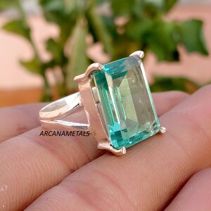 Puede incluir: Un anillo de plata con una gran piedra preciosa rectangular de color azul claro con corte esmeralda, sujeta por cuatro garras. La banda tiene un diseño dividido y el texto ARCANAMETALS es visible en el lateral del engaste metálico.