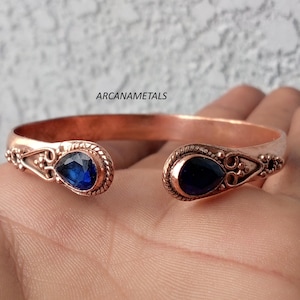 Puede incluir: Una pulsera de cobre tipo brazalete con dos piedras azul oscuro en forma de lágrima en los extremos abiertos. El metal presenta texturas intrincadas similares a cuerdas y pequeñas cuentas decorativas alrededor de cada piedra. Texto ARCANAMETALS visible.
