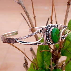 Può includere: Un bracciale a fascia color argento con una pietra ovale di colore verde brillante incastonata al centro. La fascia in metallo presenta intricati motivi a volute in rilievo e un bordo perlato attorno alla pietra, con un design aperto.