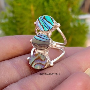 May include: A silver ring featuring three stacked oval abalone shell cabochons. The top stone displays vibrant teal and blue stripes, the middle stone shows muted grey and purple tones, and the bottom stone exhibits iridescent brown and green hues.