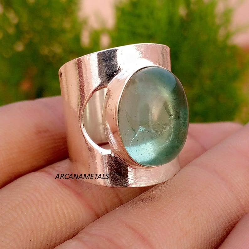 May include: A wide band ring featuring a polished metallic finish and a large, oval-shaped, translucent light green gemstone set in a bezel. The text ARCANAMETALS is printed on the side of the band. The ring has a smooth, reflective surface.