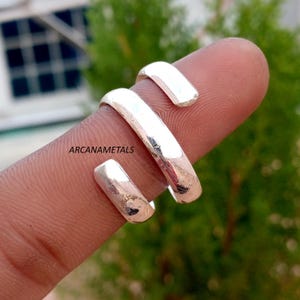 May include: A silver-coloured bypass ring with a polished, reflective surface. The ring features an open, adjustable design with two rounded ends that overlap slightly. The text ARCANAMETALS is printed in black on the surface of the metal band.