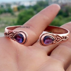 May include: A copper cuff bracelet featuring two teardrop-shaped purple amethyst gemstones at the open ends. The metal band displays intricate scrollwork and rope-style detailing. The text ARCANAMETALS is printed on the surface of the bracelet.