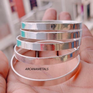 May include: A stack of five polished silver-toned bangles with a smooth, flat surface. The text ARCANAMETALS is printed in black on the bottom bangle. Each bangle features a consistent width and a reflective metallic finish throughout the set.