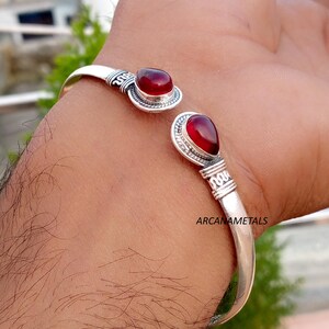 Puede incluir: Una pulsera de plata tipo brazalete con dos piedras preciosas rojas en forma de lágrima en los extremos abiertos. La banda de metal muestra detalles intrincados tipo cuerda y patrones grabados cerca de las piedras. El texto ARCANAMETALS es visible.