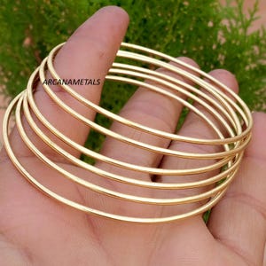 Può includere: Un set di cinque bracciali rigidi circolari, sottili e lisci, realizzati in metallo color oro. I bracciali presentano una superficie lucida e riflettente con un design semplice e privo di decorazioni. Il testo ARCANAMETALS è visibile su uno.
