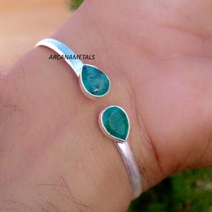 May include: A silver open cuff bracelet featuring two teardrop-shaped green emerald gemstones at each end. The metal band has a smooth, polished finish. The text ARCANAMETALS is printed in black near the upper gemstone on the silver band.
