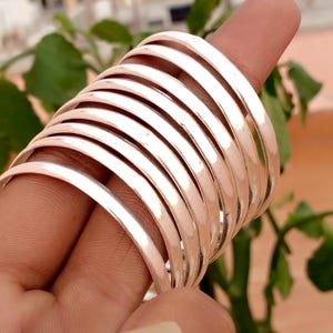 May include: A set of ten thin, polished silver-colored metal bangles stacked together. Each bangle features a smooth, rounded surface with a reflective finish. The bracelets are arranged in a parallel formation, creating a uniform appearance.