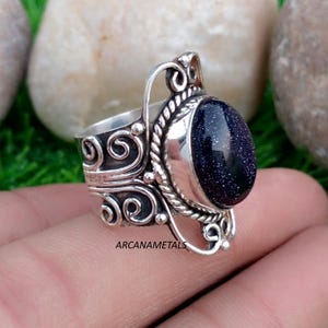 May include: A silver ring featuring a large, oval-shaped dark blue sandstone cabochon with sparkling inclusions. The metal band is decorated with intricate silver scrollwork and a twisted rope border surrounding the stone. The text ARCANAMETALS is visible.