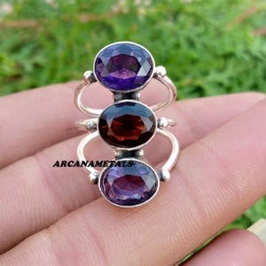 May include: A silver ring featuring three vertically aligned oval faceted gemstones. The top and bottom stones are purple amethyst, while the centre stone is a deep red garnet. The metal band has a split design, and the text ARCANAMETALS is visible.