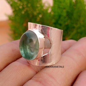 May include: A wide silver band ring featuring a polished finish and a large, smooth, oval-shaped translucent green gemstone set in a bezel. The metal band has a distinct cutout design on the side, and the text ARCANAMETALS is visible on the surface.