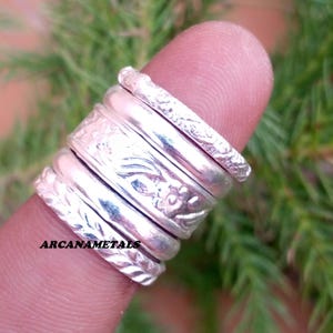 May include: A stack of five silver rings with varying textures and patterns, including floral engravings and braided designs. The text ARCANAMETALS is visible on the bottom ring. The rings are arranged vertically on a finger.