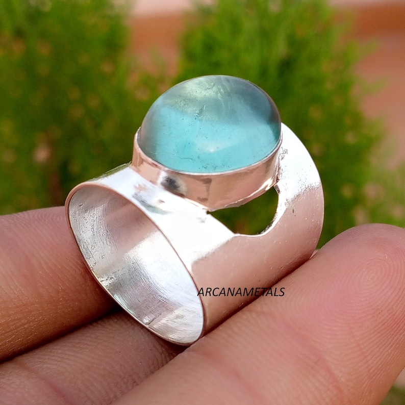 May include: A wide silver band ring featuring a smooth, polished finish and a large, round, translucent light blue cabochon stone set in a raised bezel. The text ARCANAMETALS is clearly visible on the side of the silver band surface.