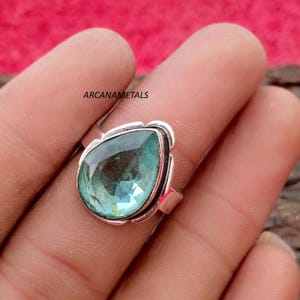 以下が含まれることがあります： 磨かれた金属製のベゼルにセットされた、大きな洋ナシ型のファセットカットが施されたティールカラーの宝石が付いたシルバーリング。リングの上部にはARCANAMETALSという文字が見えます。宝石は半透明で、深い青緑色の色調とファセットが特徴です。