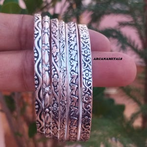 Puede incluir: Un conjunto de seis brazaletes de metal plateado con distintos patrones en relieve, incluyendo líneas onduladas, motivos florales y formas de delfines. Las pulseras están dispuestas una al lado de la otra y el texto ARCANAMETALS es visible a la derecha.