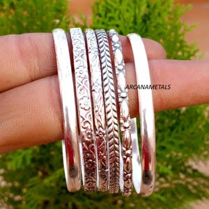 Può includere: Un set di sei bracciali rigidi in metallo color argento con vari design, tra cui superfici lisce, motivi floreali, una trama intrecciata e un motivo ovale ripetuto. Il testo ARCANAMETALS è visibile sul lato destro.