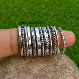 May include: A set of ten silver-toned metal rings arranged in a row, featuring various textures including twisted rope, smooth bands, beaded patterns, and engraved floral motifs. The text ARCANAMETALS is visible above the rings.