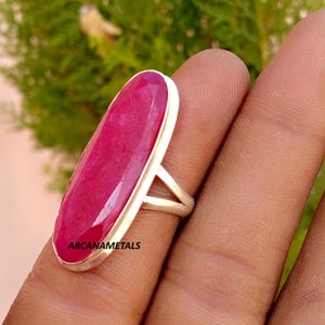 Op de afbeelding: Een zilveren ring met een grote, langwerpige, ovaal geslepen robijn gezet in een gladde metalen zetting. De edelsteen heeft een diepe magenta-rode kleur met natuurlijke insluitingen. De tekst ARCANAMETALS staat op het oppervlak van de steen.