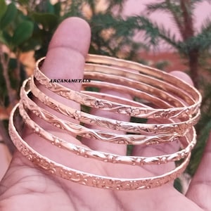Peut inclure: Un ensemble de cinq bracelets joncs en métal doré ornés de motifs floraux et ondulés en relief. Chaque bracelet circulaire présente une finition polie et une largeur uniforme. Le texte ARCANAMETALS est imprimé en noir sur le bracelet supérieur.