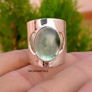 May include: A wide silver-toned band ring featuring a large, oval-shaped, translucent pale green cabochon gemstone set in the center. The metal surface has a polished finish and the text ARCANAMETALS is printed clearly on the lower portion.