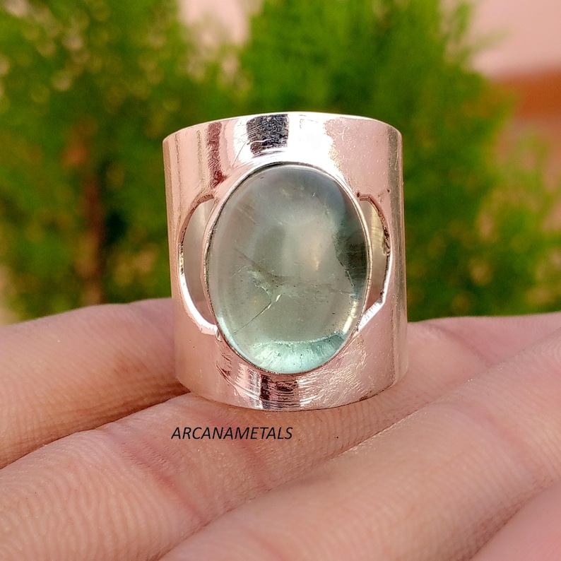May include: A wide silver-toned band ring featuring a large, oval-shaped, translucent pale green cabochon gemstone set in the center. The metal surface has a polished finish and the text ARCANAMETALS is printed clearly on the lower portion.