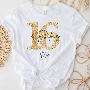 Könnte beinhalten: Ein weißes kurzärmeliges T-Shirt mit einer großen goldfarbenen Glitzergrafik der Zahl 16. Der Text & Fabulous ist in schwarzer Schreibschrift über die Mitte der Zahlen gedruckt, darunter steht der Name Mia ebenfalls in schwarzer Schrift.