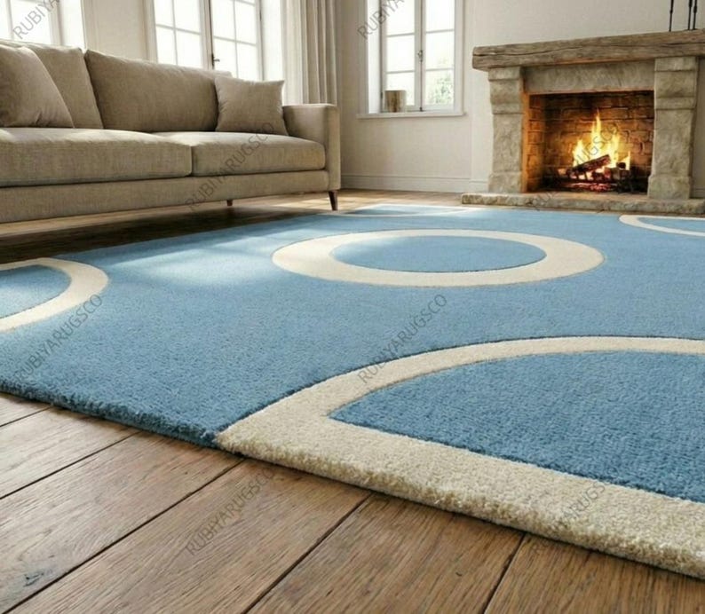 May include: A light blue area rug with a circular design in off-white. The rug is placed on a hardwood floor, with a sofa and fireplace in the background. The rug's design adds a modern touch to the room.