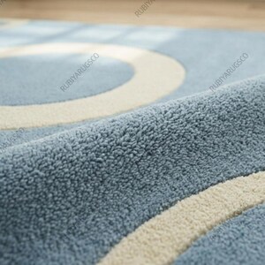 May include: Close-up of a blue rug with a cream-colored circular pattern. The rug has a soft, textured surface. The design features overlapping circles, creating a modern aesthetic. The rug's color palette is a mix of light blue and cream.