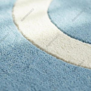 May include: Close-up of a blue and white rug with a curved design. The rug has a soft, textured surface with a light blue background and a thick, off-white curved border. The brand name "RUBIYARUGSCO" is printed repeatedly.