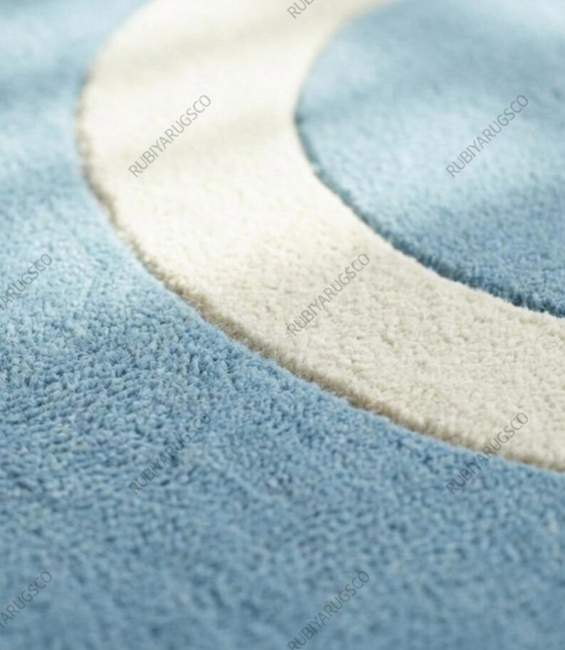 May include: Close-up of a blue and white rug with a curved design. The rug has a soft, textured surface with a light blue background and a thick, off-white curved border. The brand name "RUBIYARUGSCO" is printed repeatedly.